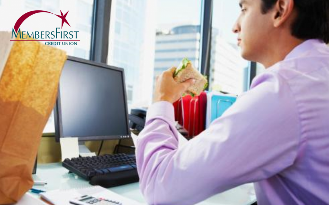 man in purple shirt sitting at desk in computer chair in front of computer in large office building eating sandwich out of brown paper sack