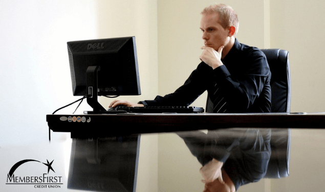 Caucasian man sitting at large desk in front of computer in black button up shirt with reflection on shiny surface of desk