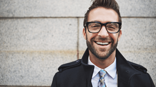 Caucasian man with brown hair in black glasses wearing coat and collared shirt with tie smiling